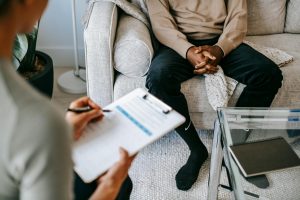 Man sitting on a couch with hands clasped during a mental health consultation, while a healthcare professional takes notes on a clipboard.