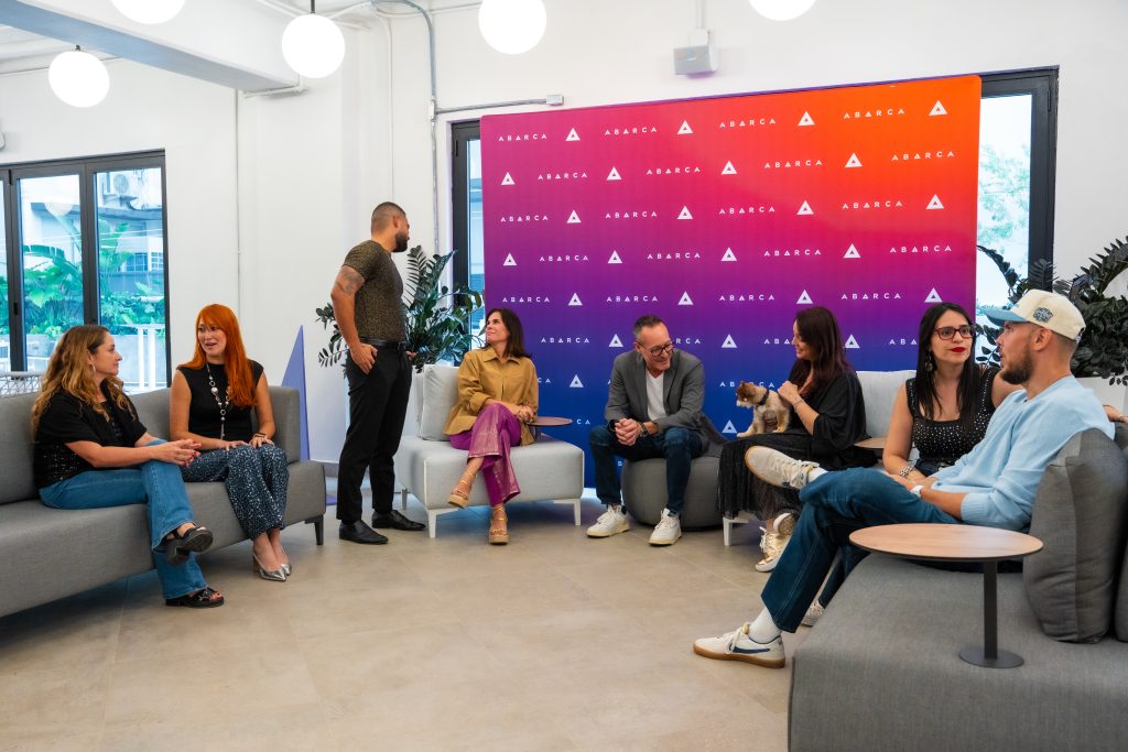 Abarca team members sitting and chatting in a bright lounge area, with a colorful Abarca-branded backdrop in the center and one person holding a small dog.