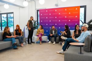 Abarca team members sitting and chatting in a bright lounge area, with a colorful Abarca-branded backdrop in the center and one person holding a small dog.