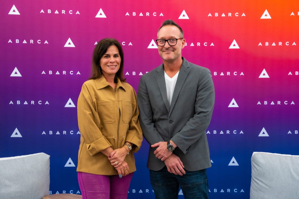 Two Abarca leaders standing and smiling in front of a colorful purple-to-orange Abarca-branded backdrop in an office lounge.