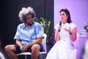 Two panelists, Mercibel Gonzalez, Executive Vice President and Chief People Officer, and Devin Baptiste of Endeavor, seated on stage during an AmplifyHER event. Mercy speaks into a microphone while Devin listens, engaged in a discussion on leadership and mentorship.