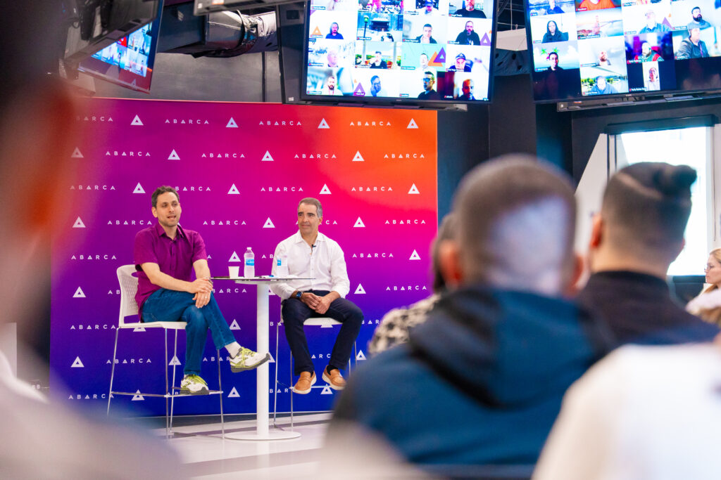 Carlos M. García and Jason Borschow seated on stage during Oasis Connections Season 4, Episode 1, speaking to an in-person audience with virtual attendees displayed on screens above them.
