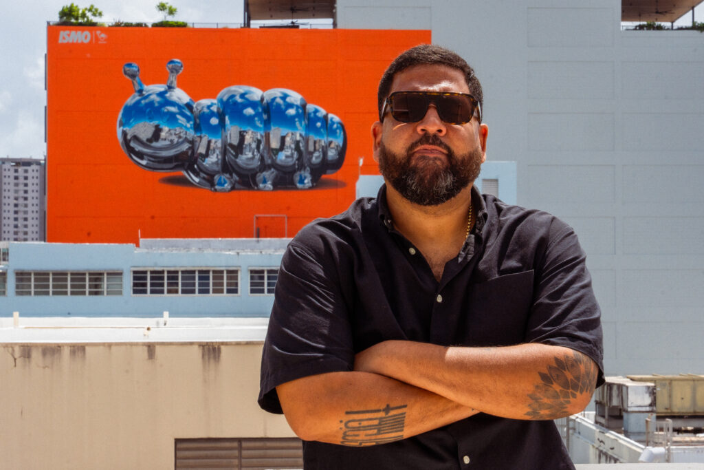 Puerto Rican artist Bikismo stands on a rooftop with his chrome caterpillar mural, The Caterpillar, on Abarca Health’s Santurce façade behind him.